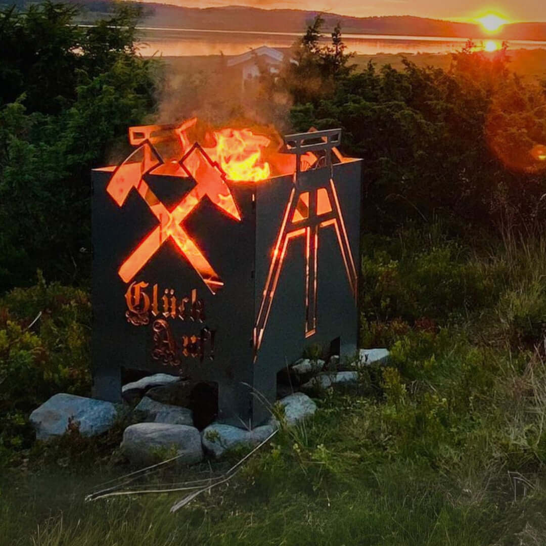 ein Feuerkorb mit Bergbaumotiven auf der Wiese eines Berges. Im Hintergrund ist der Sonnnenuntergang zu sehen